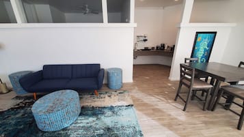 A modern living room with a dark blue sofa, matching ottoman, and a dining table with chairs. A large window provides natural light, and a ceiling fan is visible.