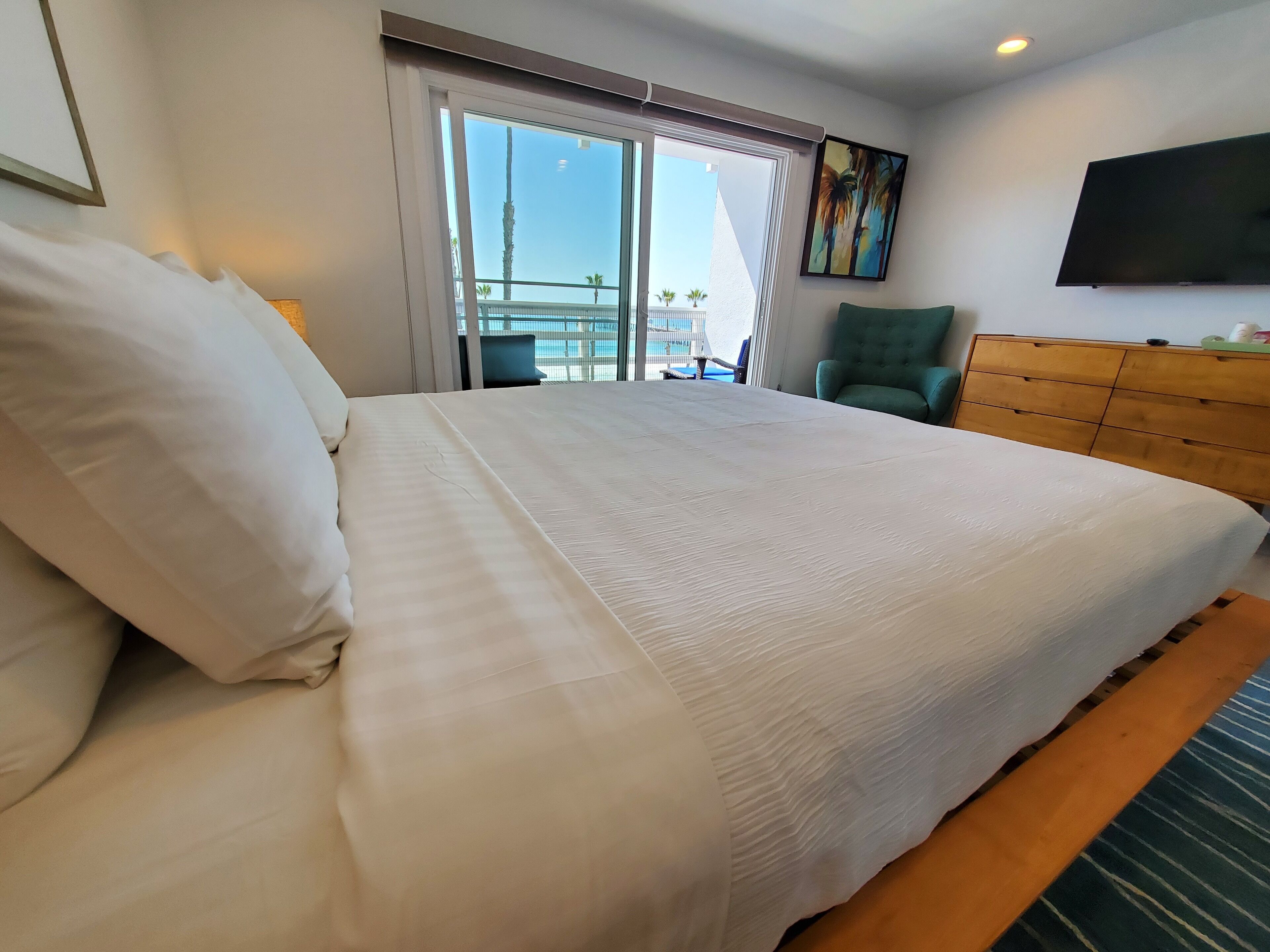 Une chambre d’hôtel avec un grand lit, un fauteuil vert, une commode en bois et un téléviseur fixé au mur.