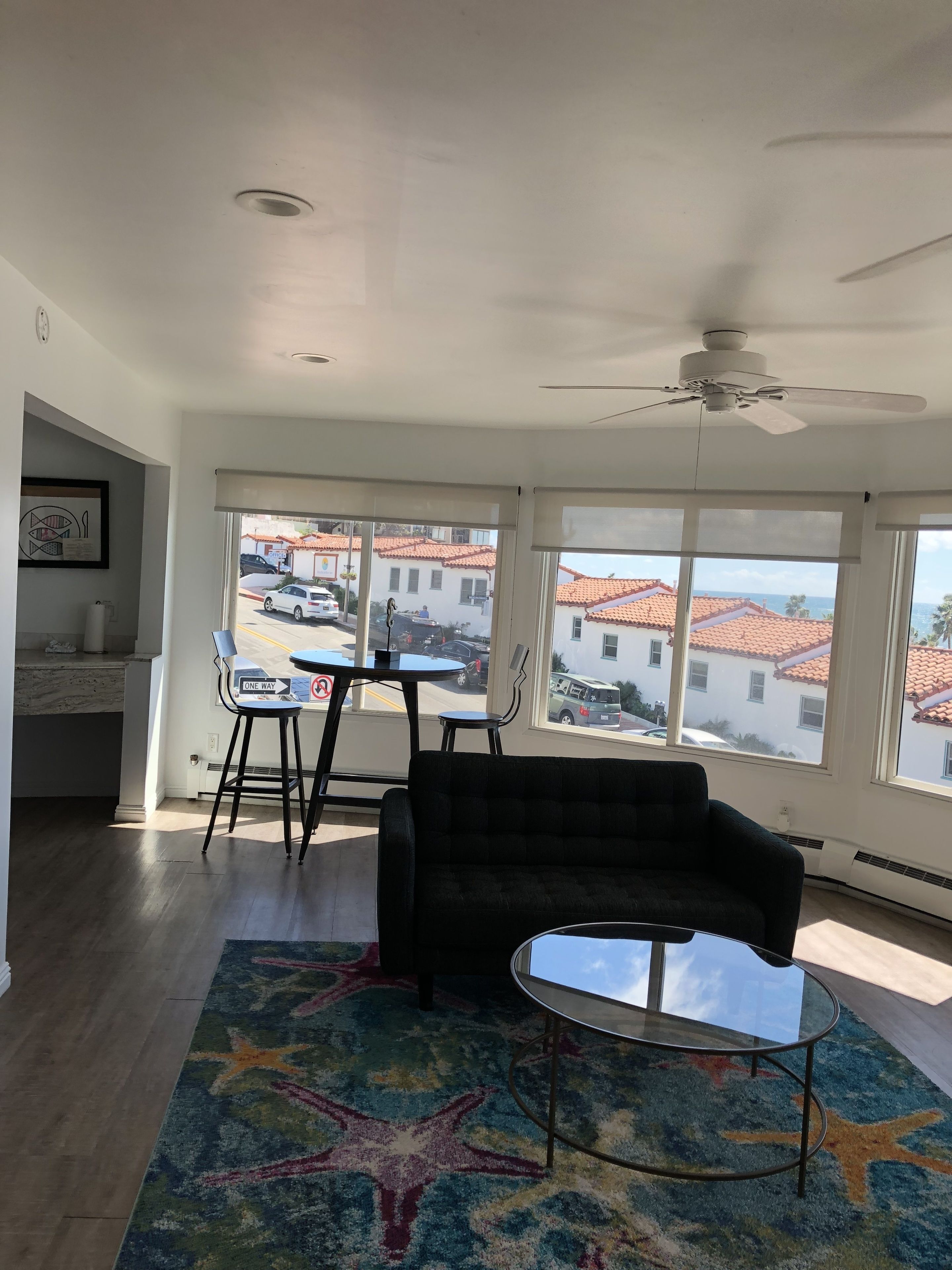 A spacious living room with a large window, a ceiling fan, a black sofa, a glass coffee table, and a colorful starfish rug.