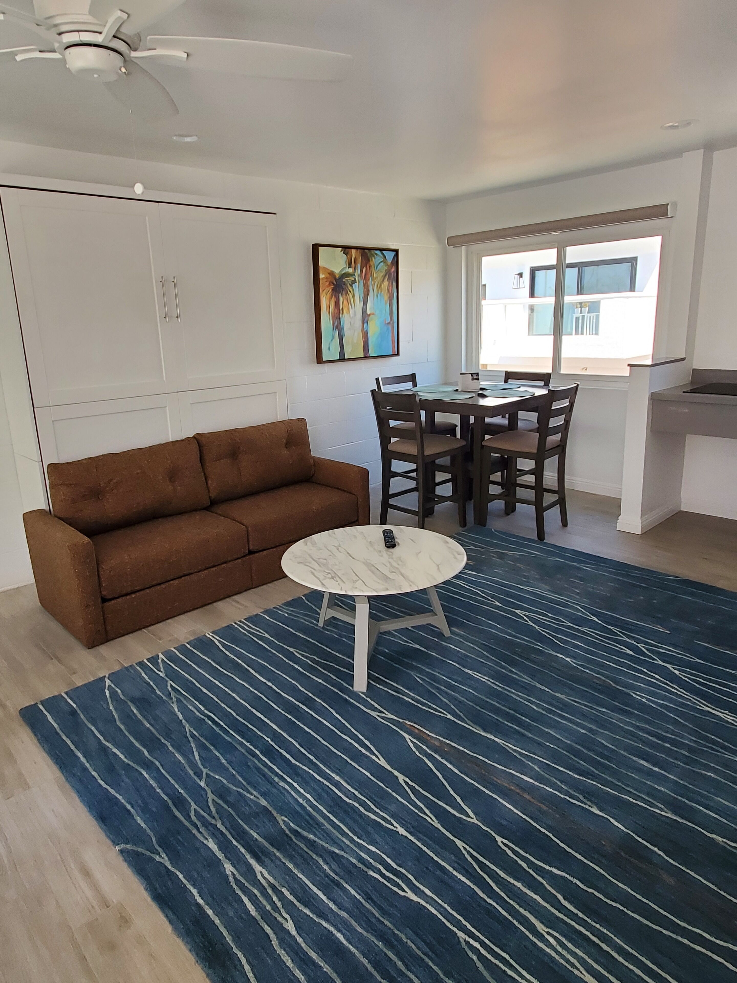 Un salon avec un canapé marron, une table basse ronde et une table à manger avec des chaises. Il y a un tableau au mur et une fenêtre donnant sur l’extérieur.