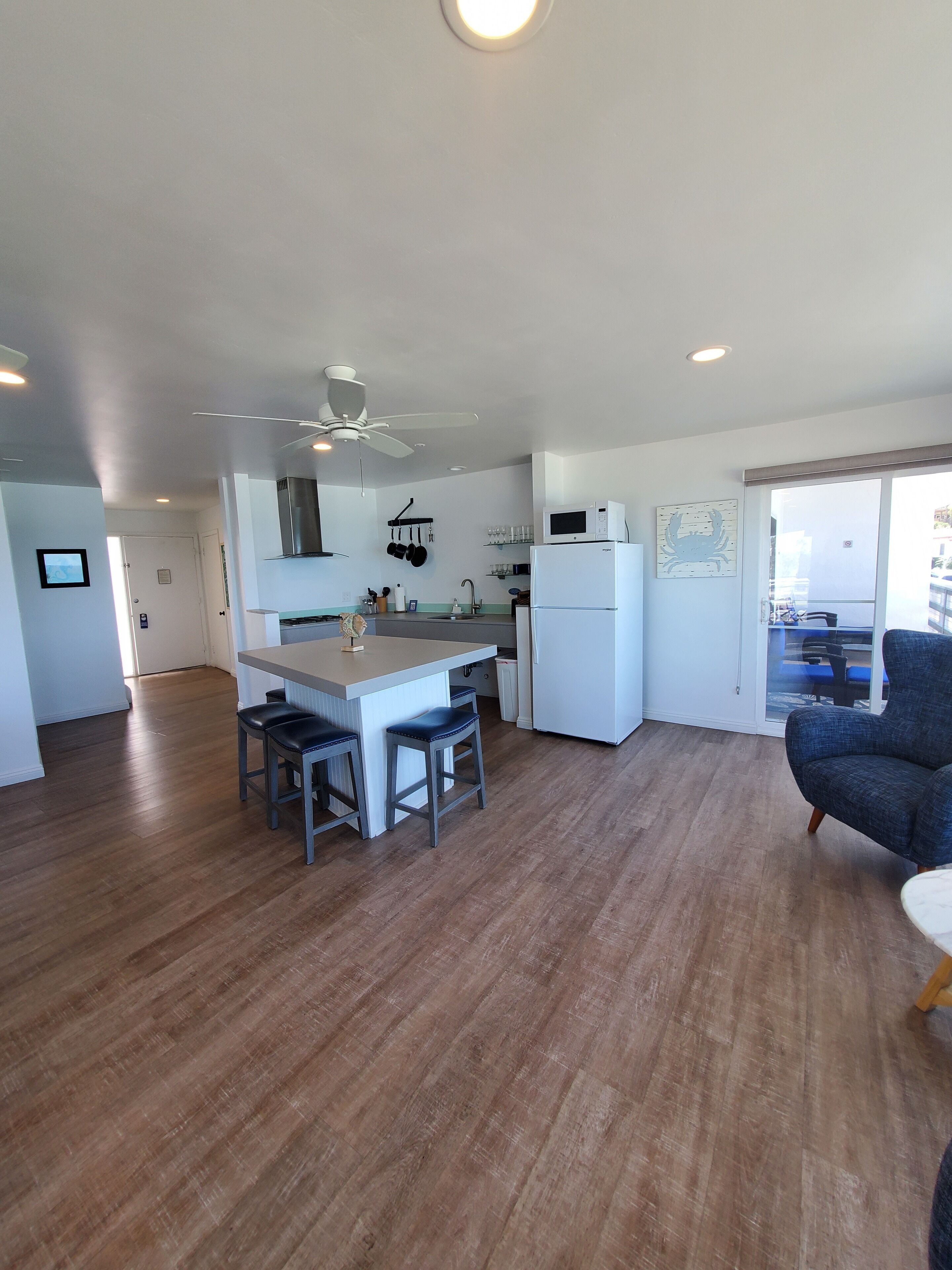 Una cocina moderna con una isla central, taburetes de bar, ventilador de techo y un comedor con una silla.