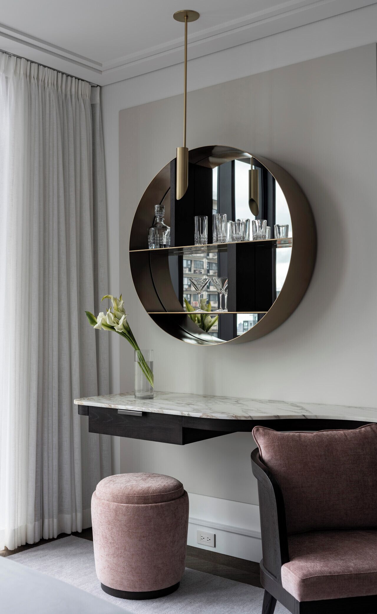 A modern interior with a round mirror, a marble-top table, a pink ottoman, and a chair.