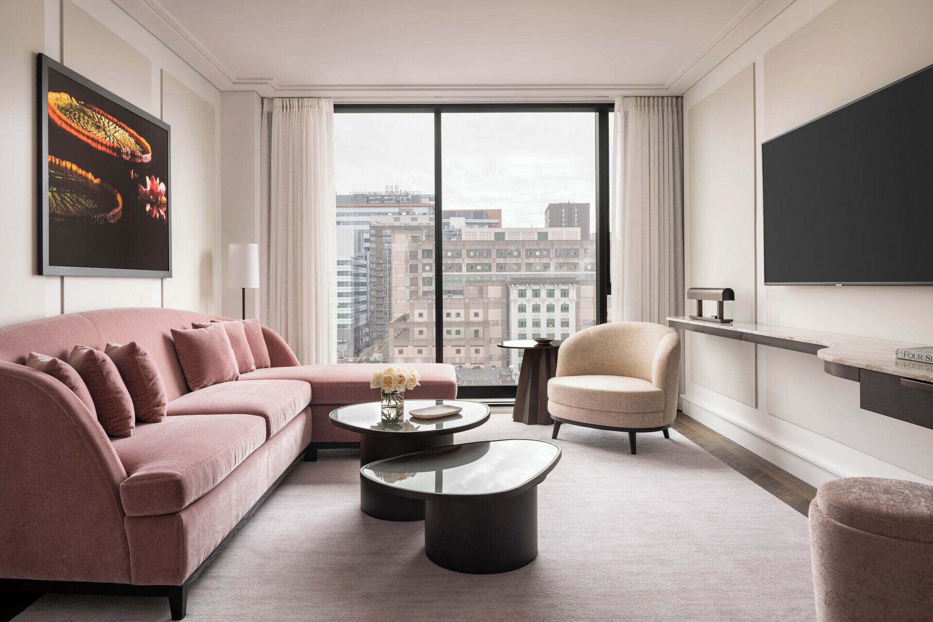 Ein modernes Hotelzimmer mit einem rosafarbenen Sofa, einem runden Couchtisch, einem beigefarbenen Sessel und einem großen Fenster mit Blick auf die Stadt.