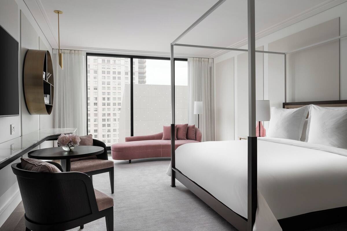 Habitación de hotel con una cama grande, un escritorio con silla, un sillón rosa y un televisor montado en la pared.