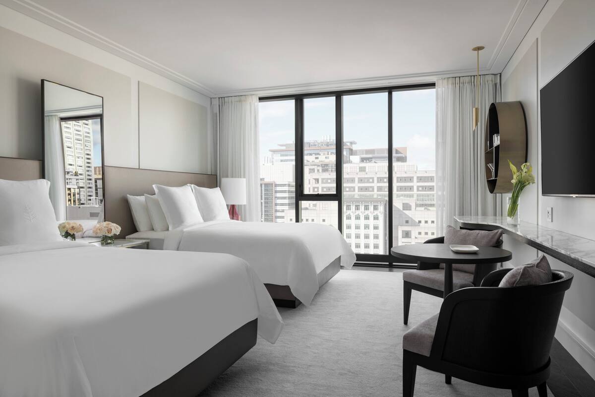Habitación de hotel con dos camas, una sala de estar con mesa y sillas, un ventanal con vistas a la ciudad y un televisor de pared.