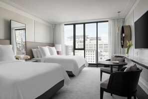 A hotel room with two beds, a sitting area with a table and chairs, a large window with a city view, and a mounted TV. - Four Seasons Hotel Montreal (Montreal)