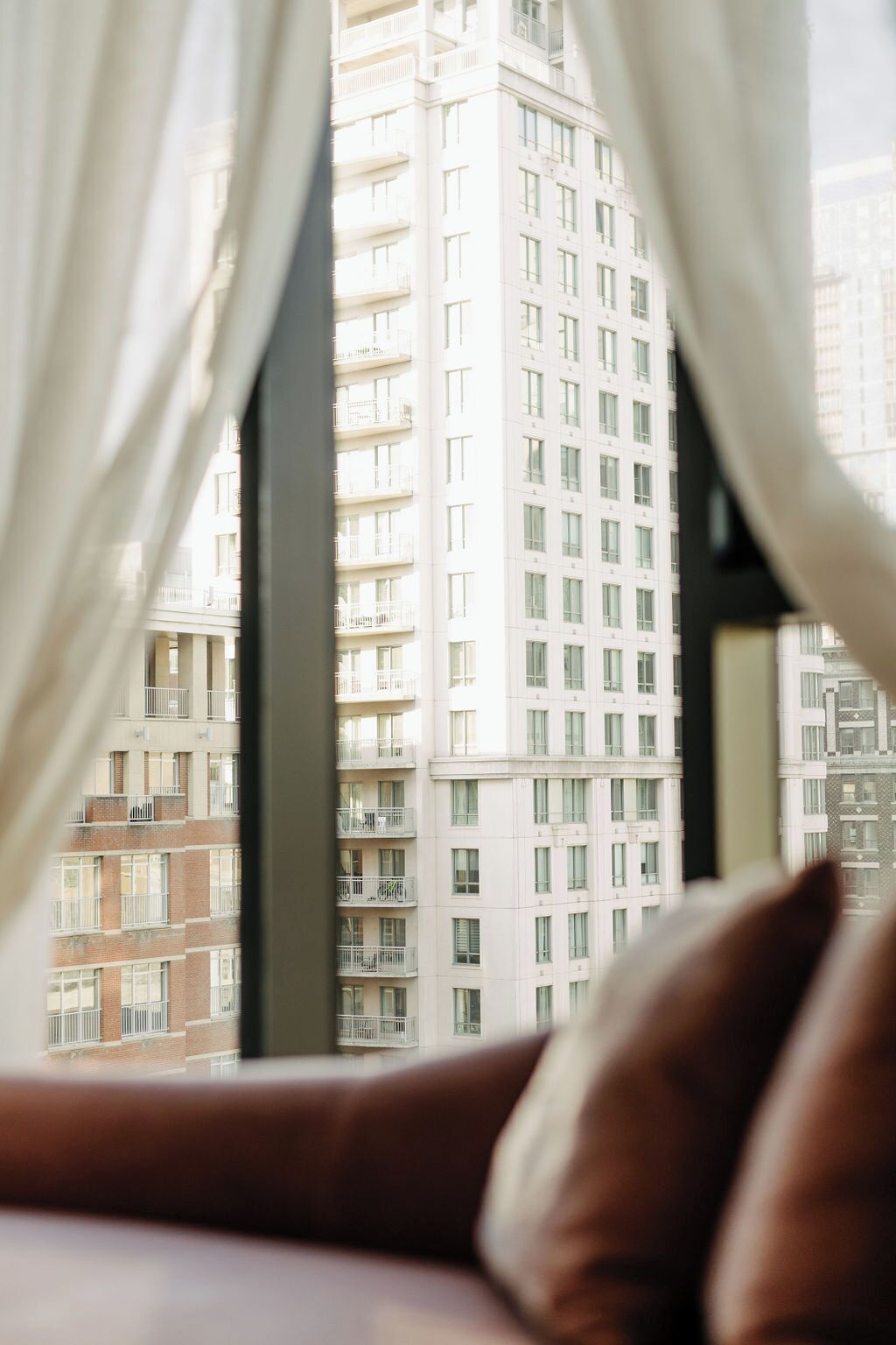 A view through a window with curtains, featuring a high-rise building with multiple balconies and windows.