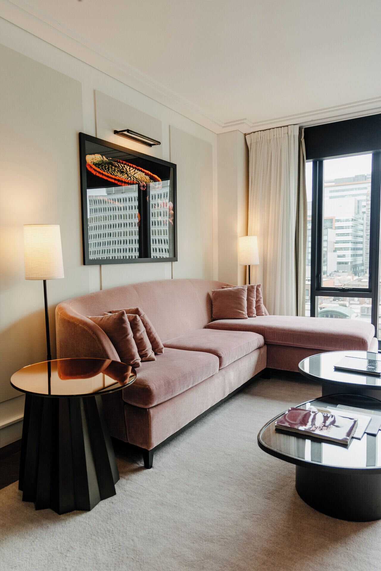 Una sala de estar moderna con un sofá seccional rosa, dos mesas auxiliares, una mesa de centro de vidrio y un ventanal con vista a la ciudad.