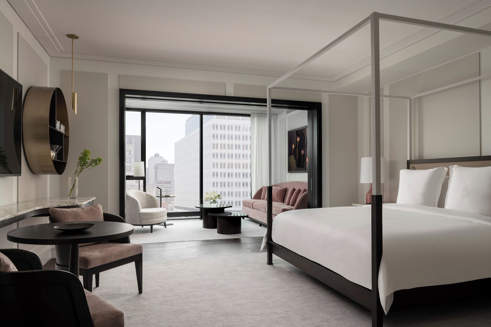 Ein modernes Hotelzimmer mit einem großen Bett, einer Sitzecke mit Sofa und Sesseln, einem runden Tisch und einem großen Fenster mit Blick auf die Stadt.