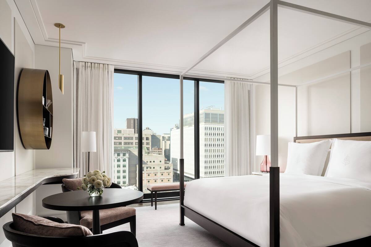 Habitación de hotel con una cama grande, una mesa redonda con un jarrón de flores, un espacio de descanso con sillas y vistas a edificios de la ciudad a través de grandes ventanales.