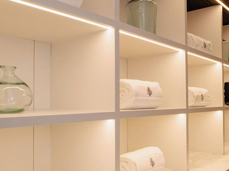 Un baño de hotel con paredes de azulejos blancos y estantes con toallas enrolladas y un logo de un árbol.