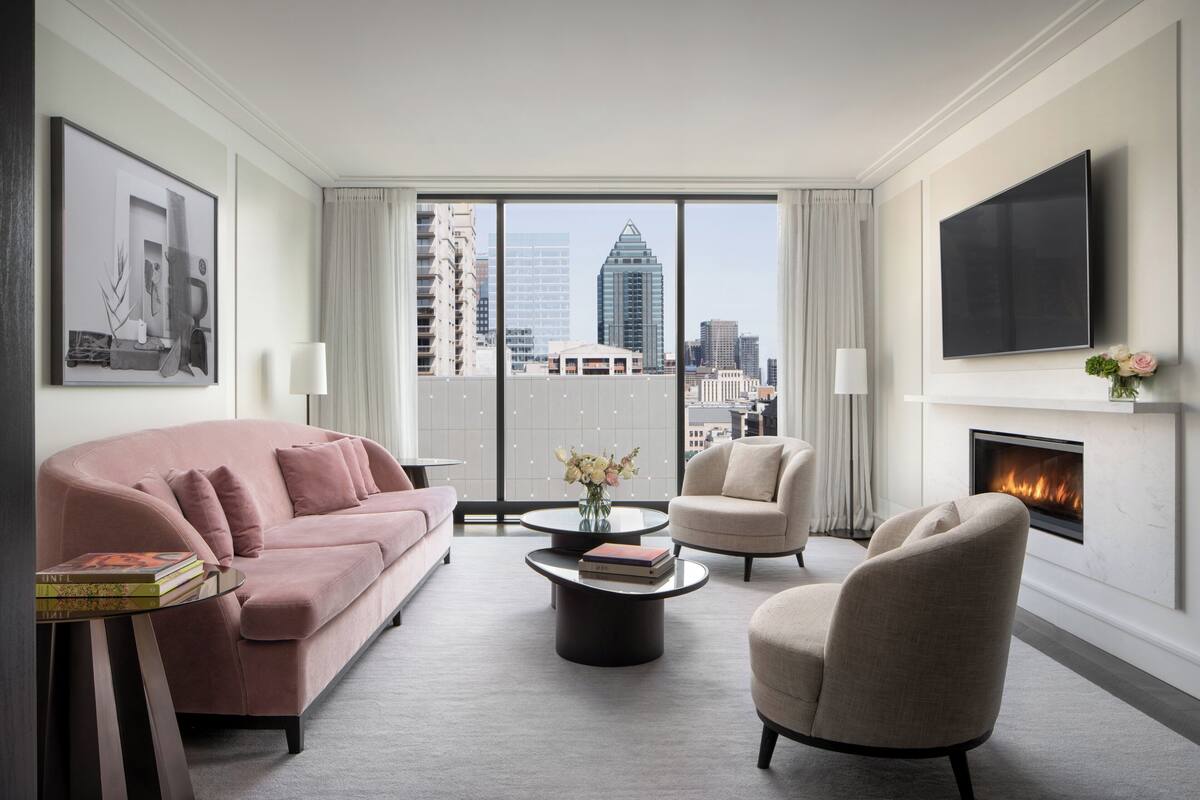 Un moderno salón con un sofá rosa, sillones beige, una mesa de centro y una chimenea. Un televisor grande está montado en la pared, y se ve una vista de la ciudad a través de las ventanas.