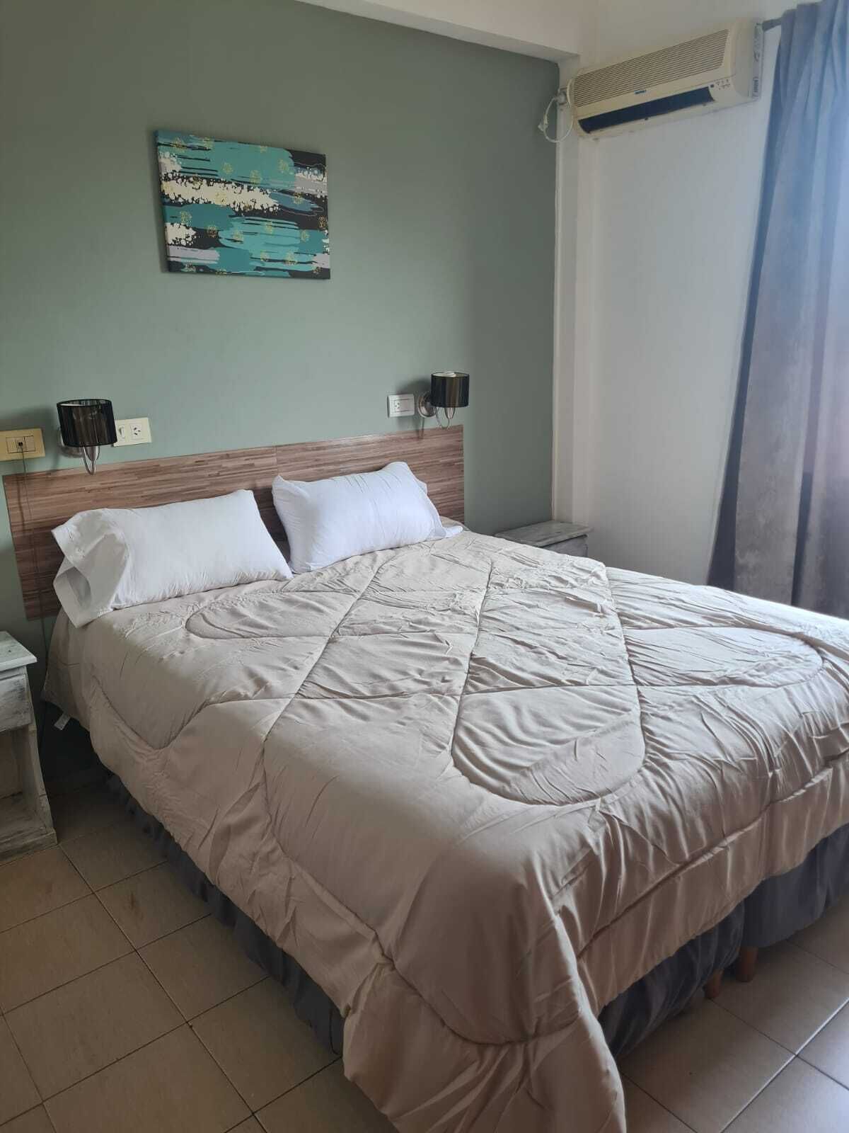 A neatly made bed with a beige quilt, two bedside lamps, a wall-mounted air conditioner, and a painting above the bed.