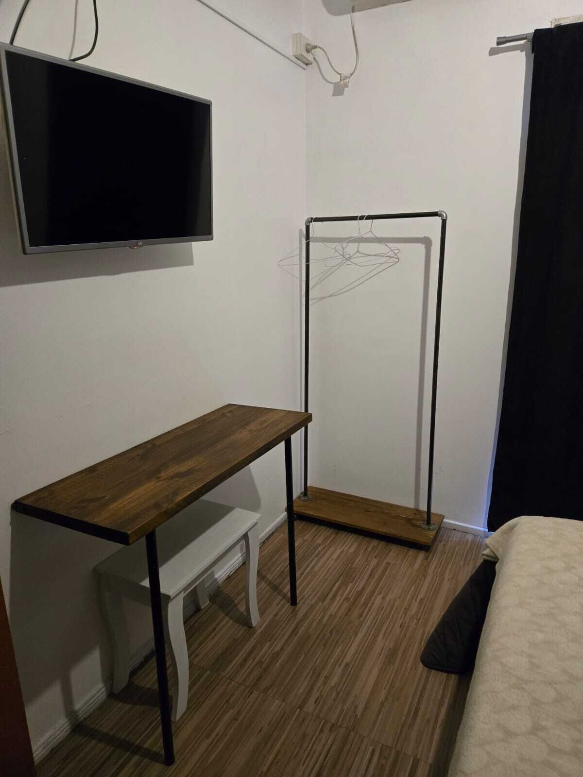 A small, functional room with a wooden desk, a white chair, a hanging TV, and a bed with a black curtain.