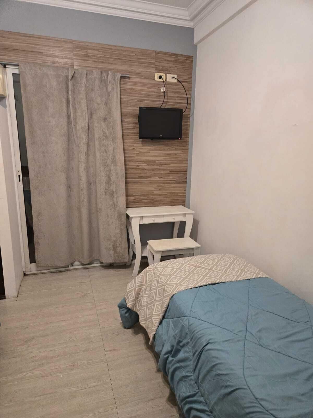A bedroom with a bed, a TV mounted on the wall, a small table, and a curtain.