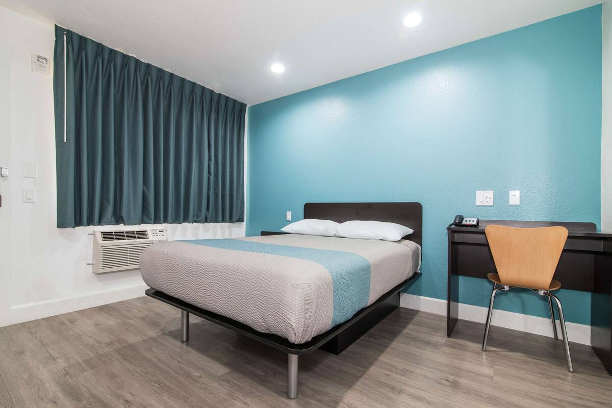 A bedroom with a bed, a desk, and a chair. There is a window with curtains, an air conditioning unit, and a wall-mounted phone.