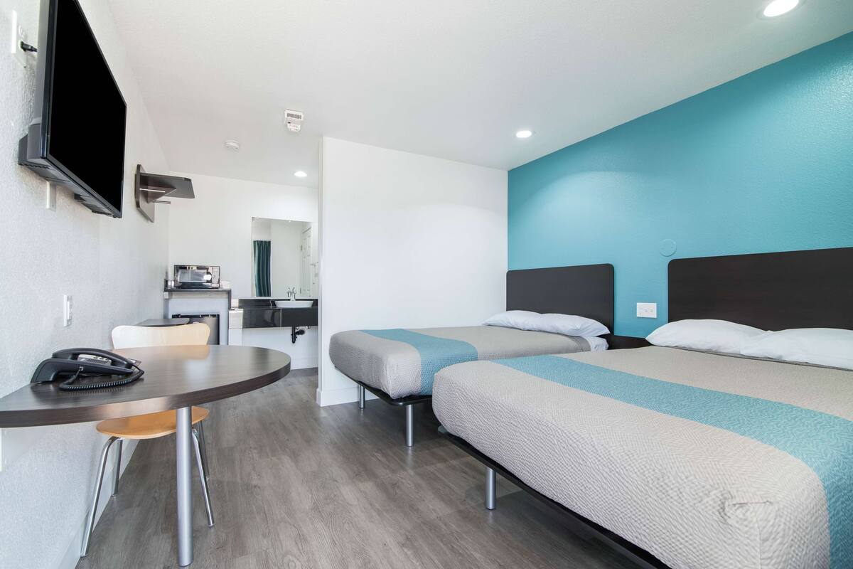 A hotel room with two beds, a desk, a chair, a TV, and a kitchenette.