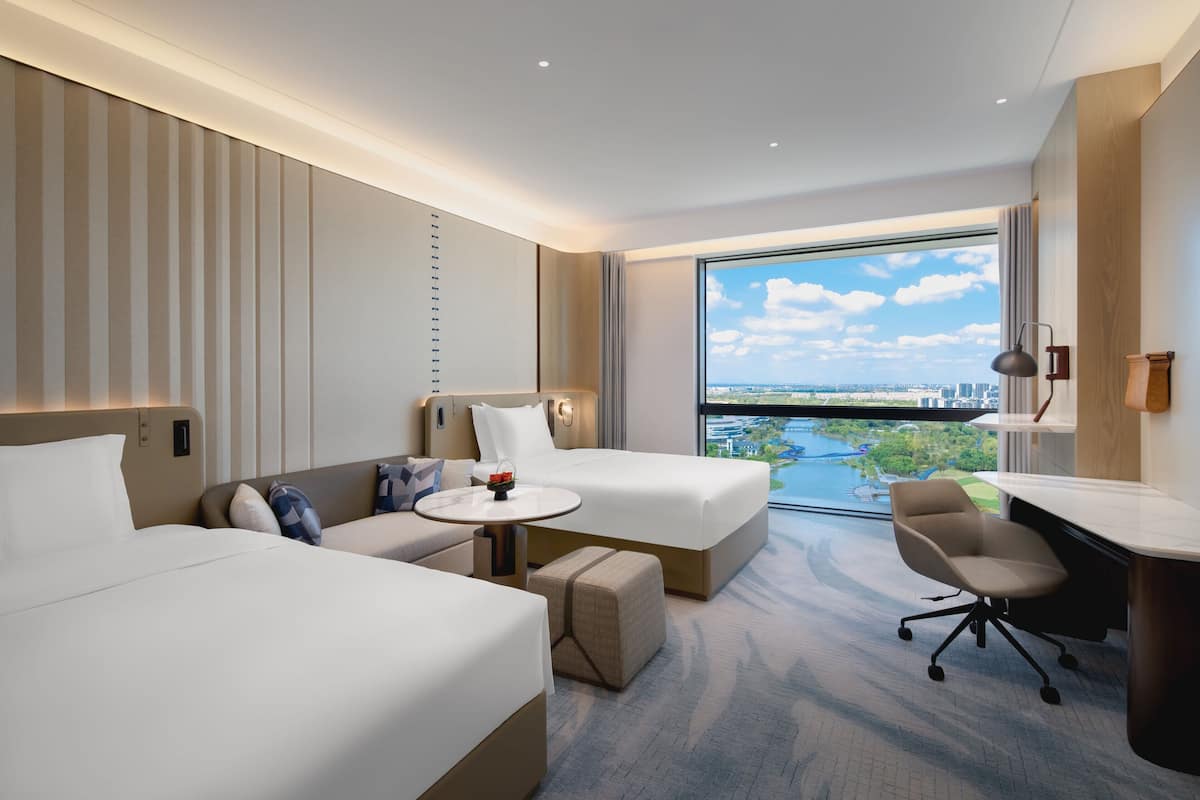 A hotel room with two beds, a small table, a desk, and a view of a cityscape and river.