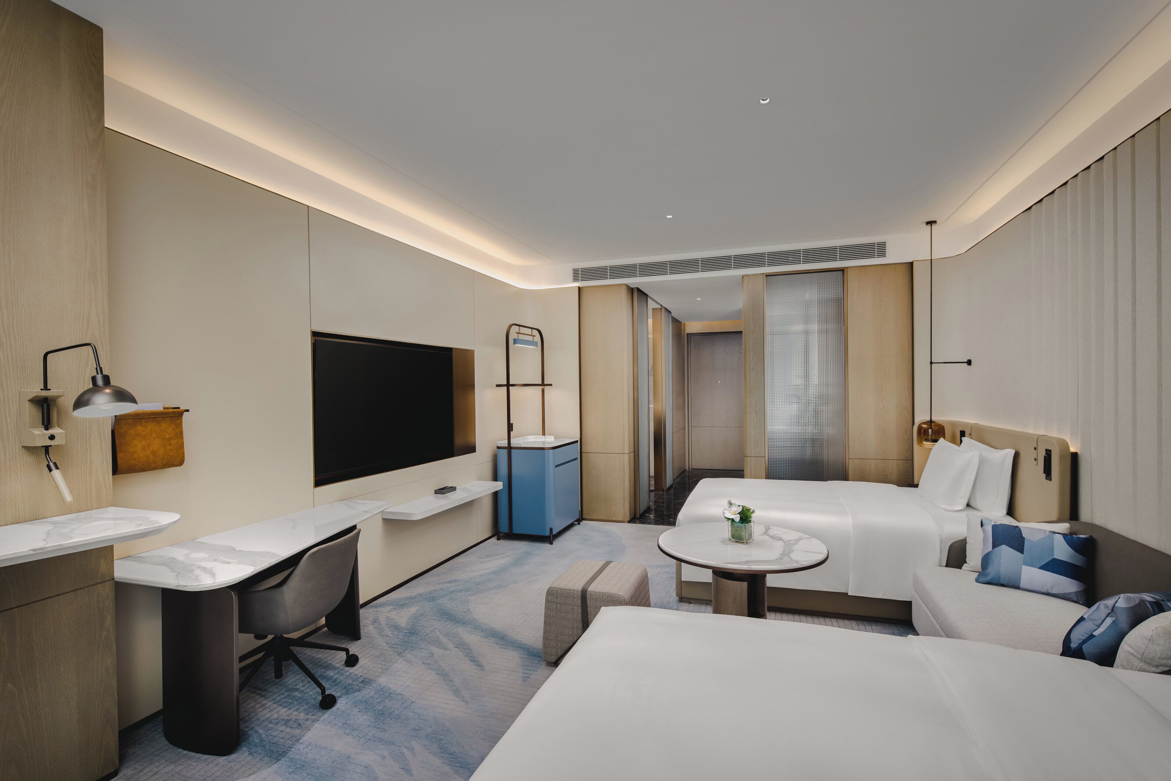 A modern hotel room with a large bed, a desk with a chair, a small table, a TV, and a bathroom with a shower and toilet.