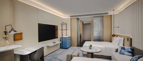 A modern hotel room with a large bed, a desk with a chair, a small table, a TV, and a bathroom with a shower and toilet.
