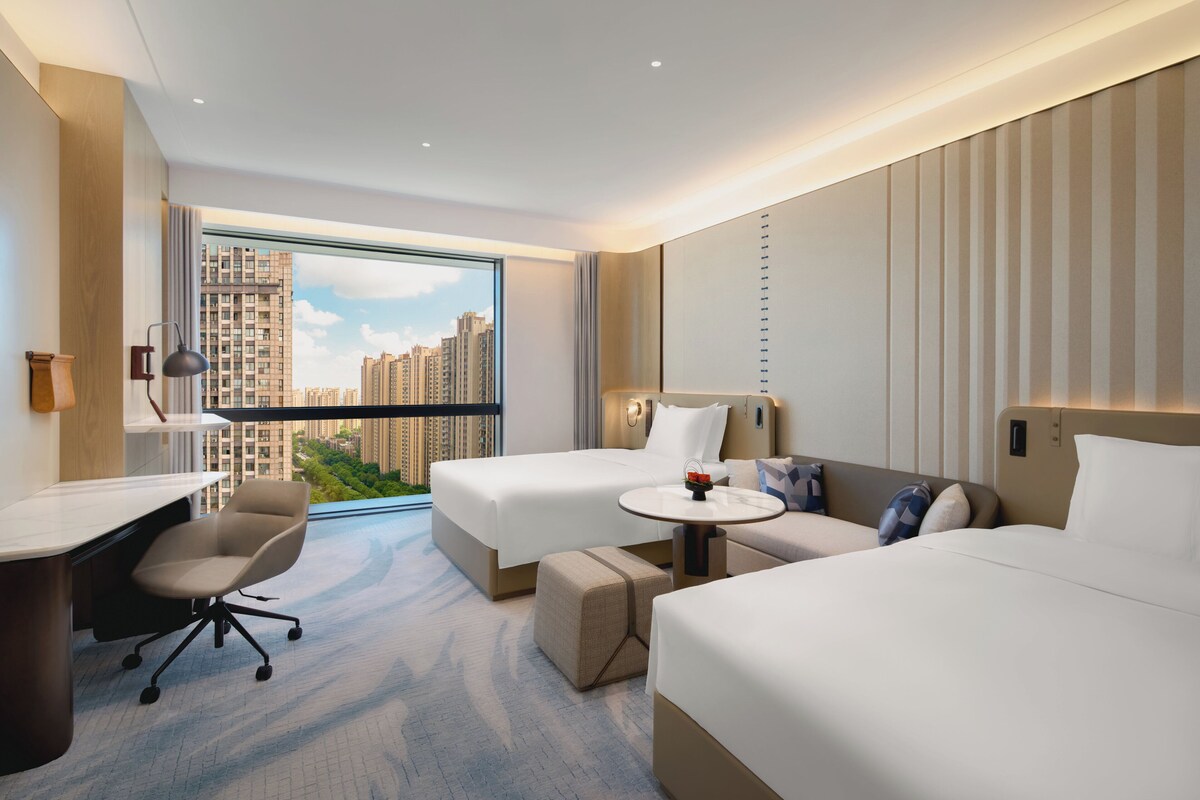 Een moderne hotelkamer met twee bedden, een bureau en uitzicht op hoge gebouwen.
