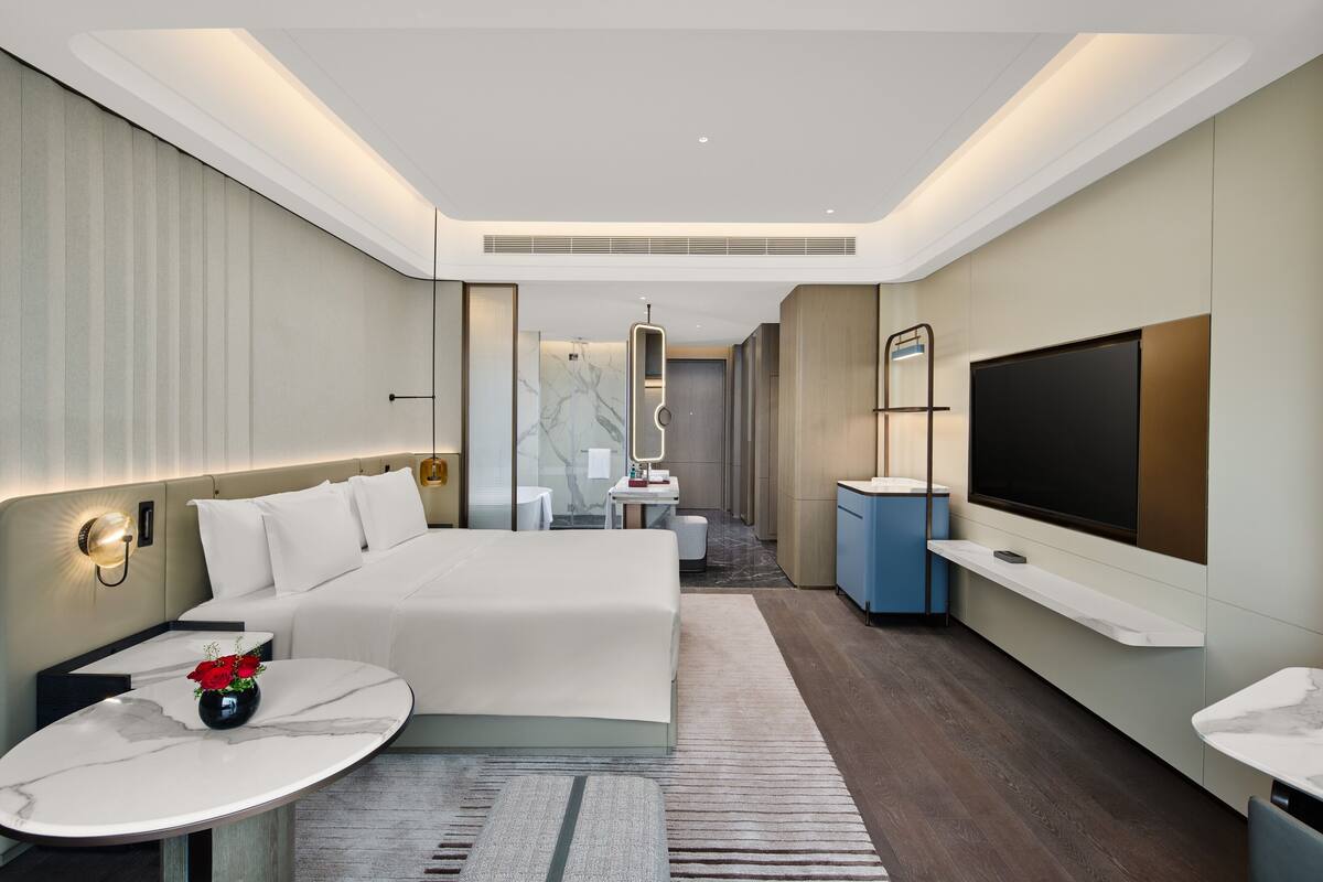 A modern hotel room with a large bed, a small desk, a TV, and a bathroom in the background.