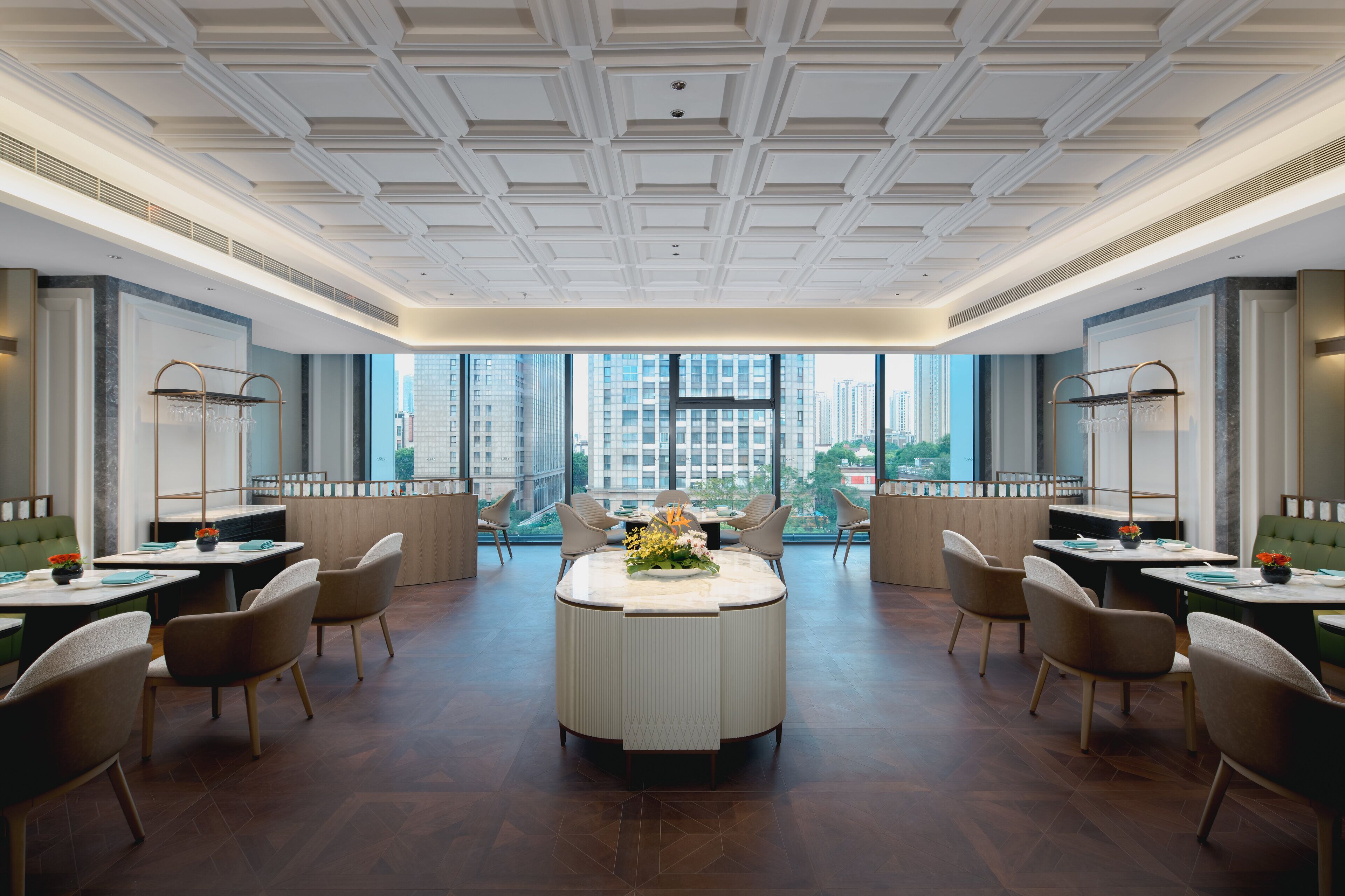 A modern hotel lobby with a central round table, multiple seating arrangements, and large windows offering city views.
