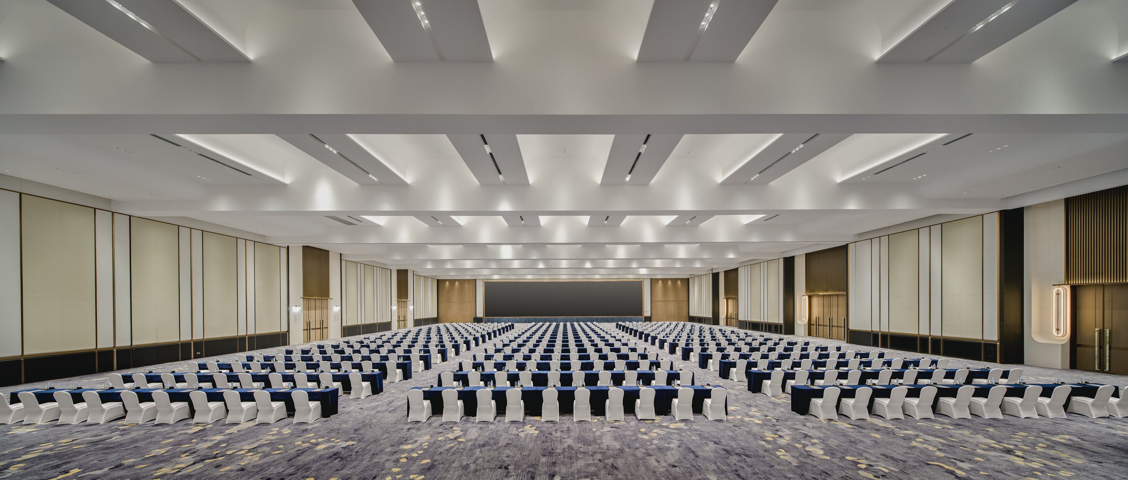 Un salón de conferencias amplio con filas de mesas y sillas, piso alfombrado y un diseño moderno de techo.