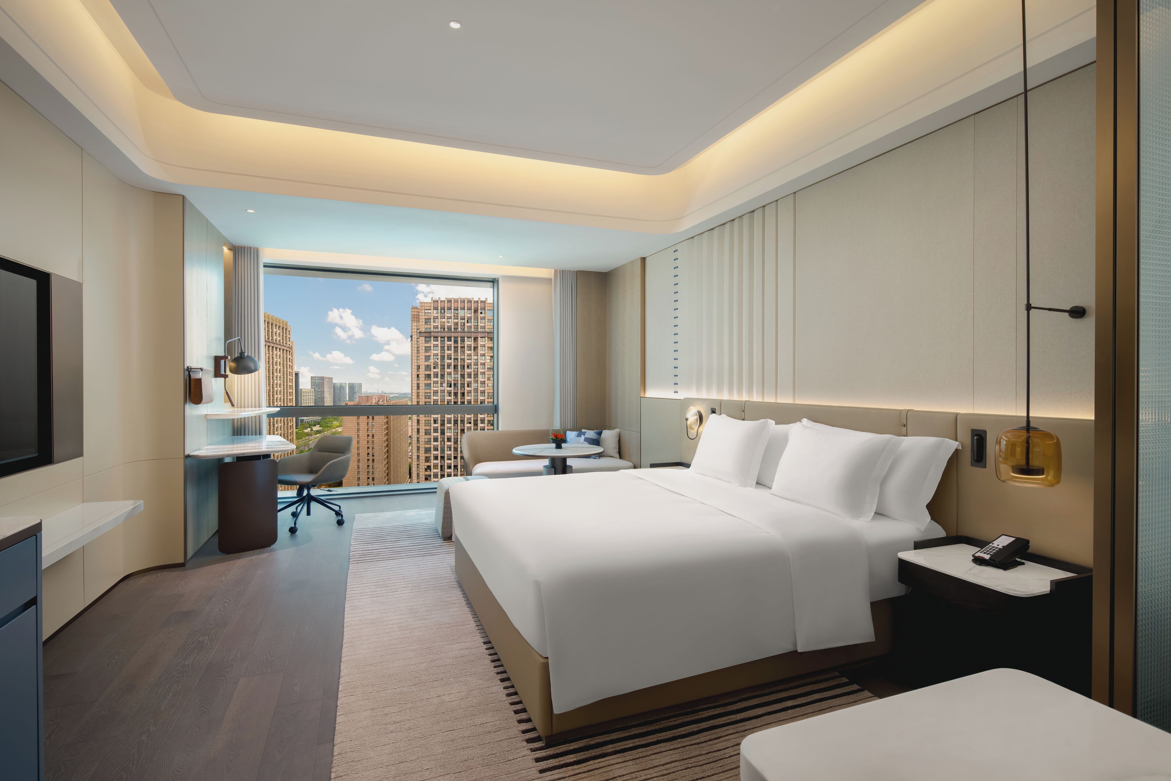 A modern hotel room with a large bed, a desk, and a view of tall buildings.
