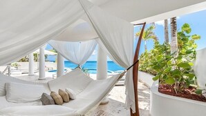 A white canopy bed with pillows, a pool, and palm trees in the background. - Villa Mumbai (Lowlands)