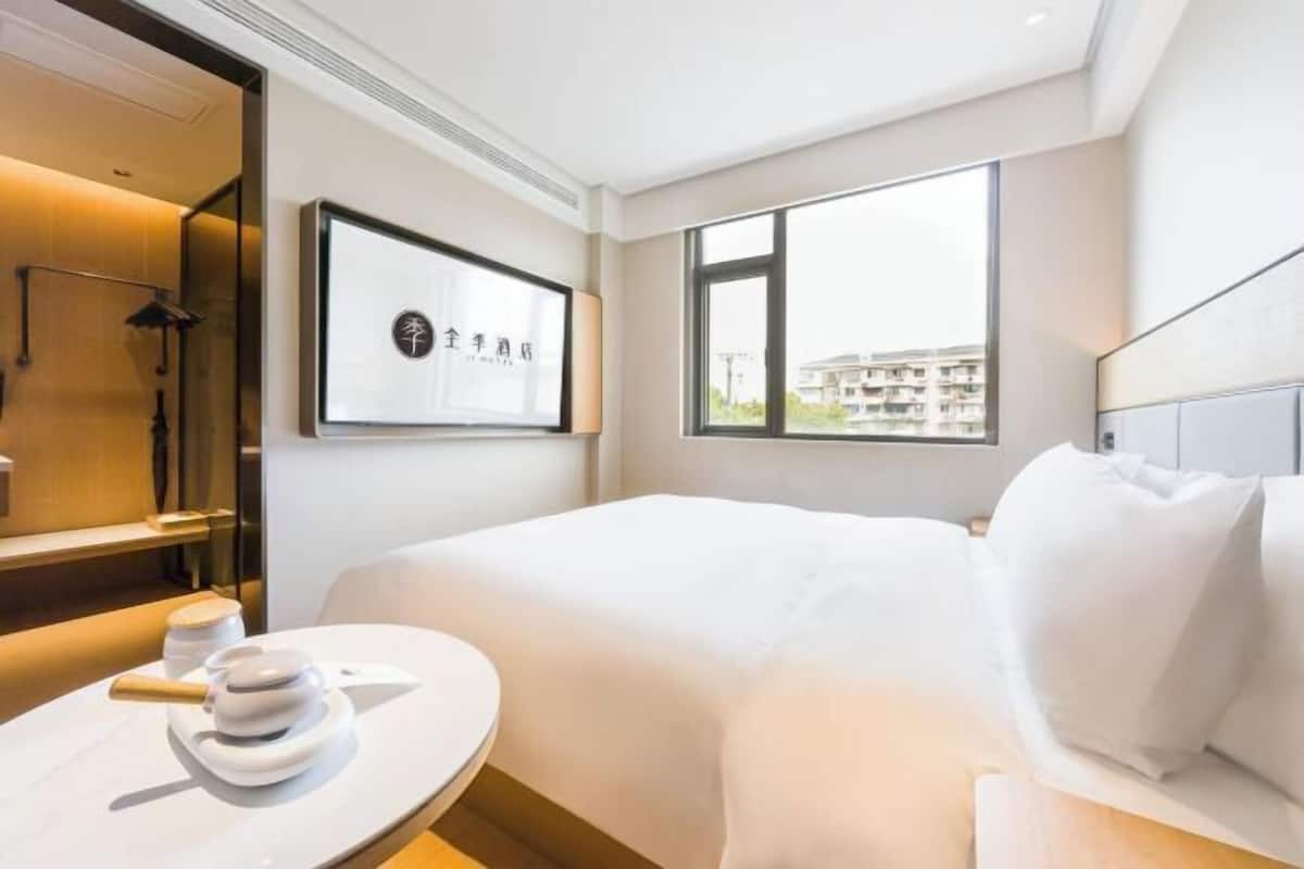 A hotel room with a bed, a round table with a white object, a framed picture on the wall, and a window with a view of buildings.
