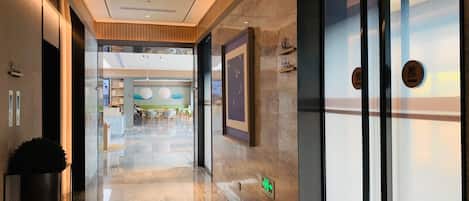 A hotel corridor with a polished marble floor, wooden paneling on the walls, and a glass door with a push button.