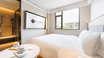 Una habitación de hotel moderna con una cama impecable, una mesa redonda con un objeto decorativo, un ventanal con vistas a edificios y una obra de arte enmarcada en la pared.