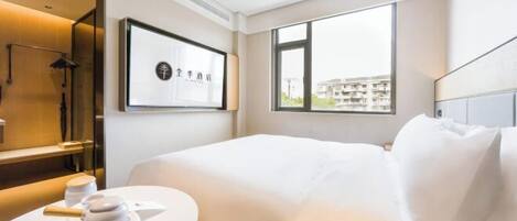 A modern hotel room with a neatly made bed, a round table with a decorative item, a large window with a view of buildings, and a framed artwork on the wall.