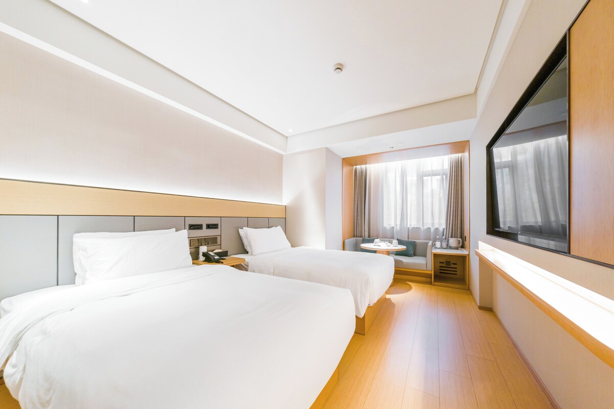Habitación de hotel con dos camas, una mesita, un sofá, una ventana con cortinas y un espejo grande.