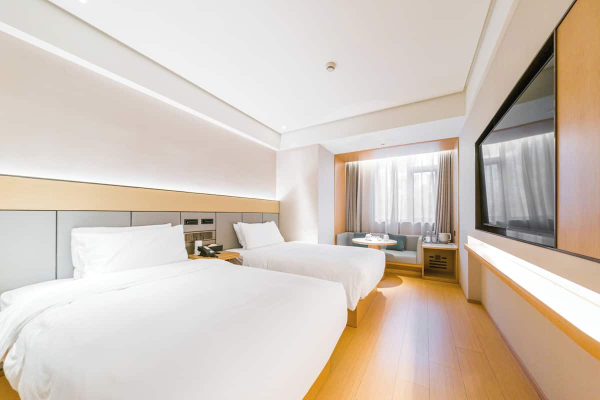 A hotel room with two beds, a small table, a sofa, a window with curtains, and a large mirror.