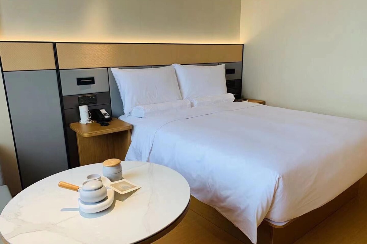 Una habitación de hotel con una cama bien tendida, una mesita de noche con una lámpara, un teléfono y una pequeña mesa con tetera y taza.