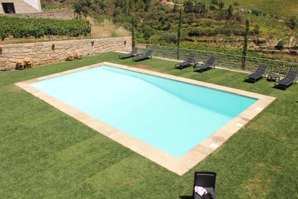 Una piscina rectangular con borde de piedra, rodeada por un césped cuidado y un muro de piedra.