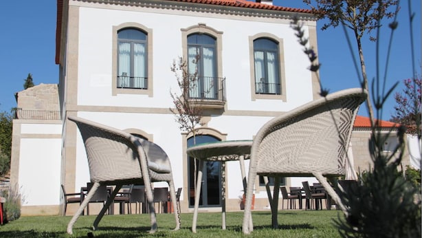 Un bâtiment à deux étages, doté d’un toit recouvert de tuiles rouges, de grandes fenêtres et d’un balcon. On aperçoit, au premier plan, des chaises et des tables en osier.