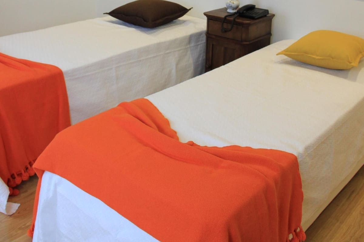 Habitación de hotel con dos camas individuales, cada una con una colcha blanca y mantas naranjas. Hay dos cuadros en la pared, una mesita de noche con una lámpara y un teléfono.