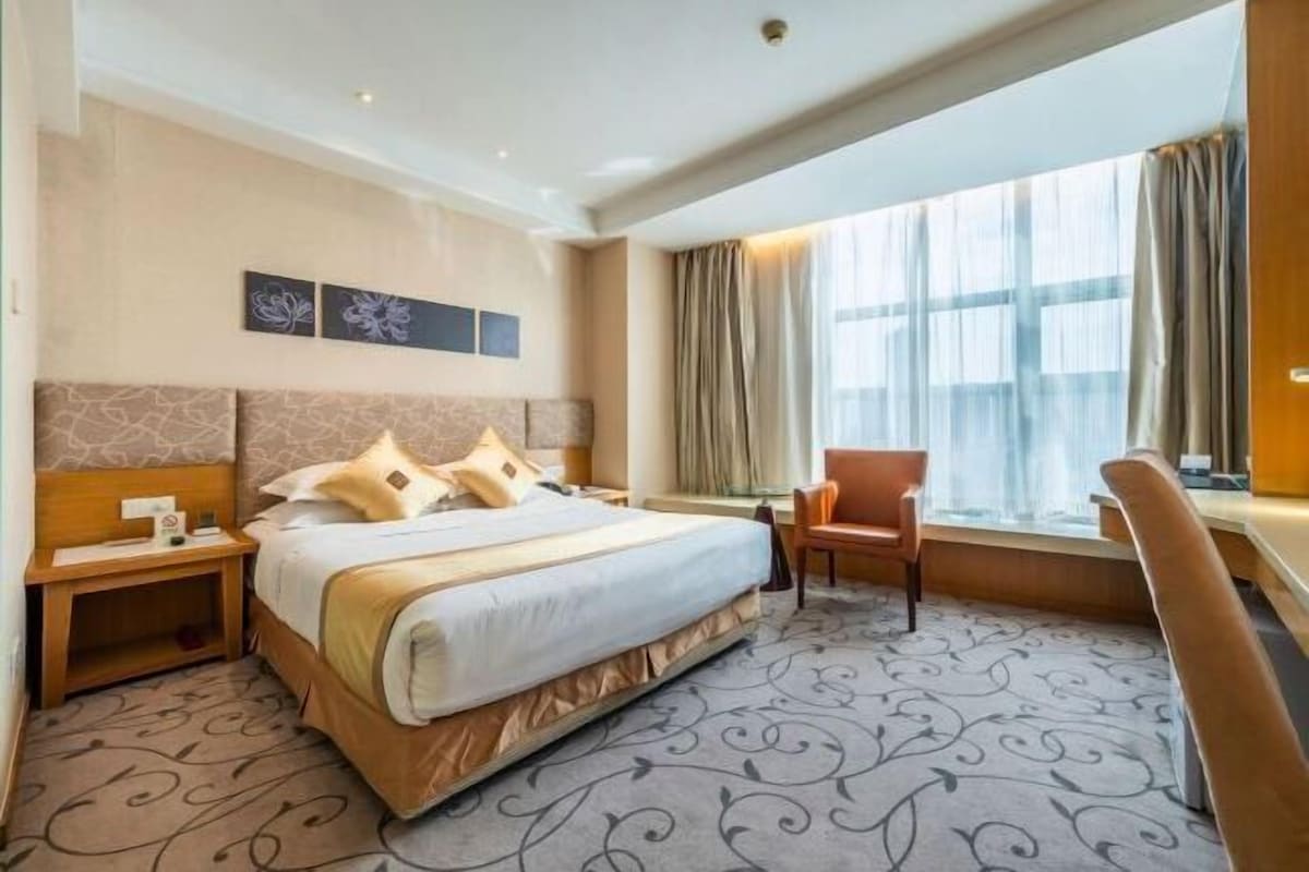 A hotel room with a large bed, a desk, and a chair. There is a window with curtains, a wall with artwork, and a carpet with a pattern.