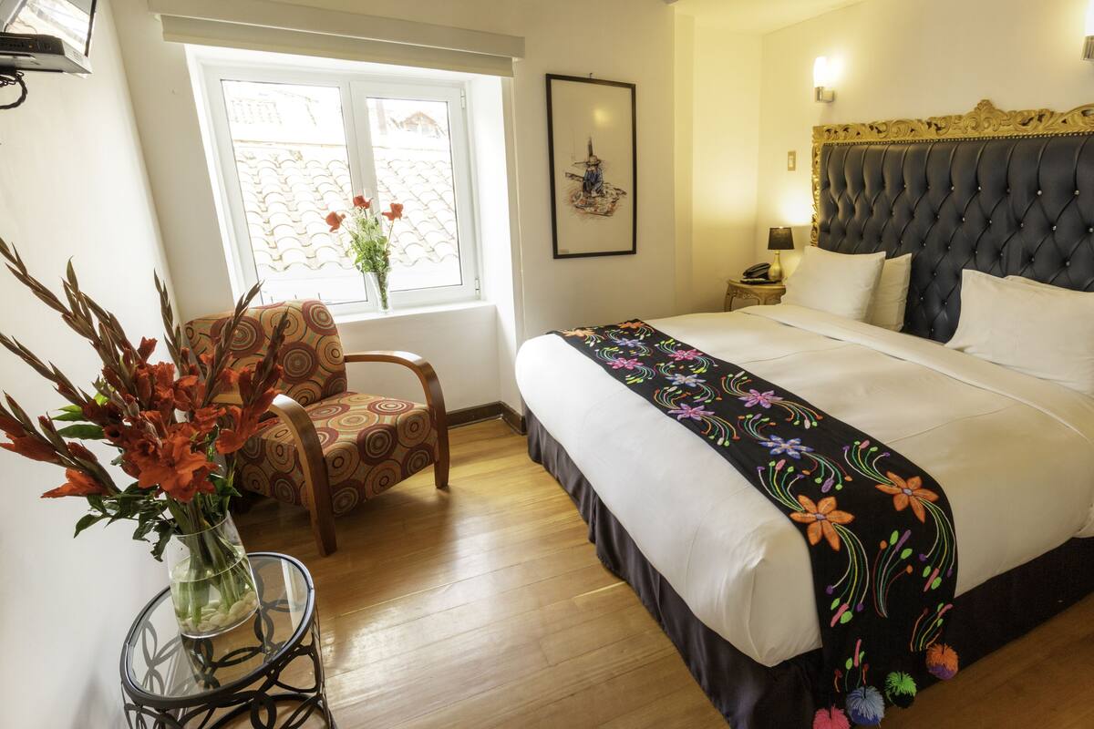 A hotel room with a large bed, a chair, a small table with a vase of flowers, and a window with a view of the outside.