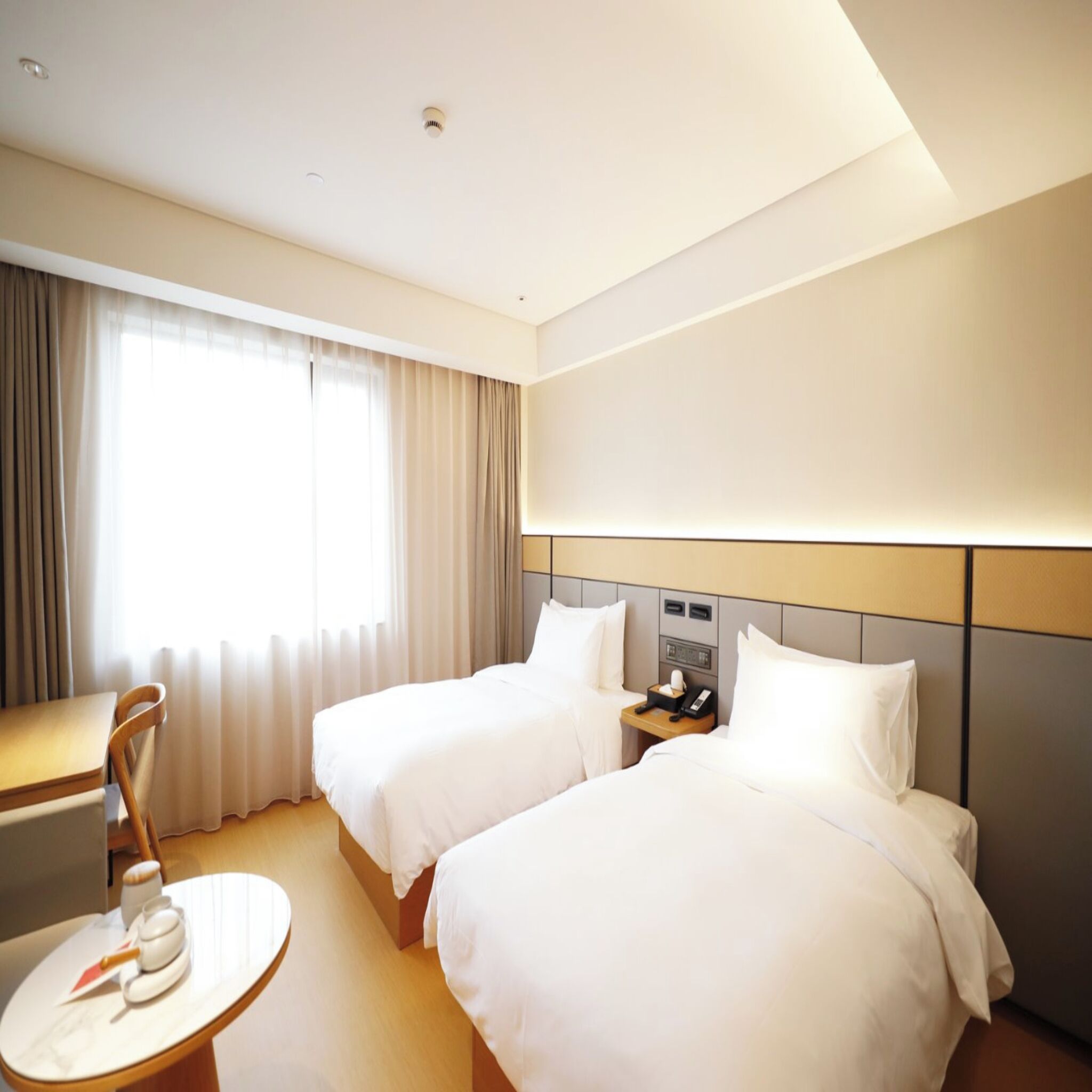 Een hotelkamer met twee bedden, een bureau, een stoel, een klein tafeltje met een waterkoker en een raam met gordijnen.