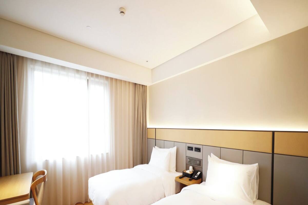 Ein Hotelzimmer mit zwei Betten, einem Schreibtisch, einem Stuhl, einem kleinen Tisch mit Teekanne und einem Fenster mit Vorhängen.
