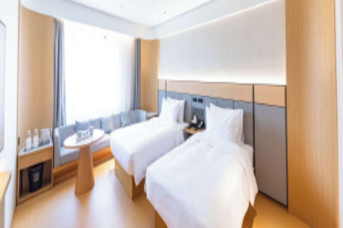 A hotel room with two beds, a small seating area, and a window providing natural light.