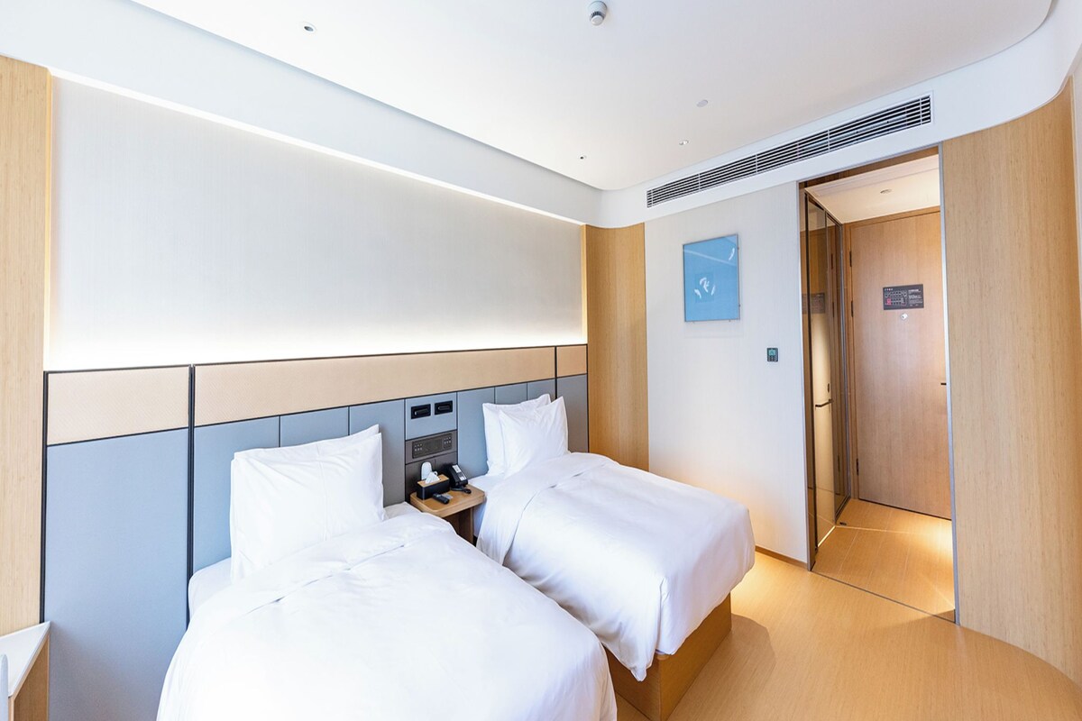 Habitación de hotel con dos camas, una mesita de noche con teléfono y lámpara, un panel de control en la pared y una puerta que da a otra habitación.