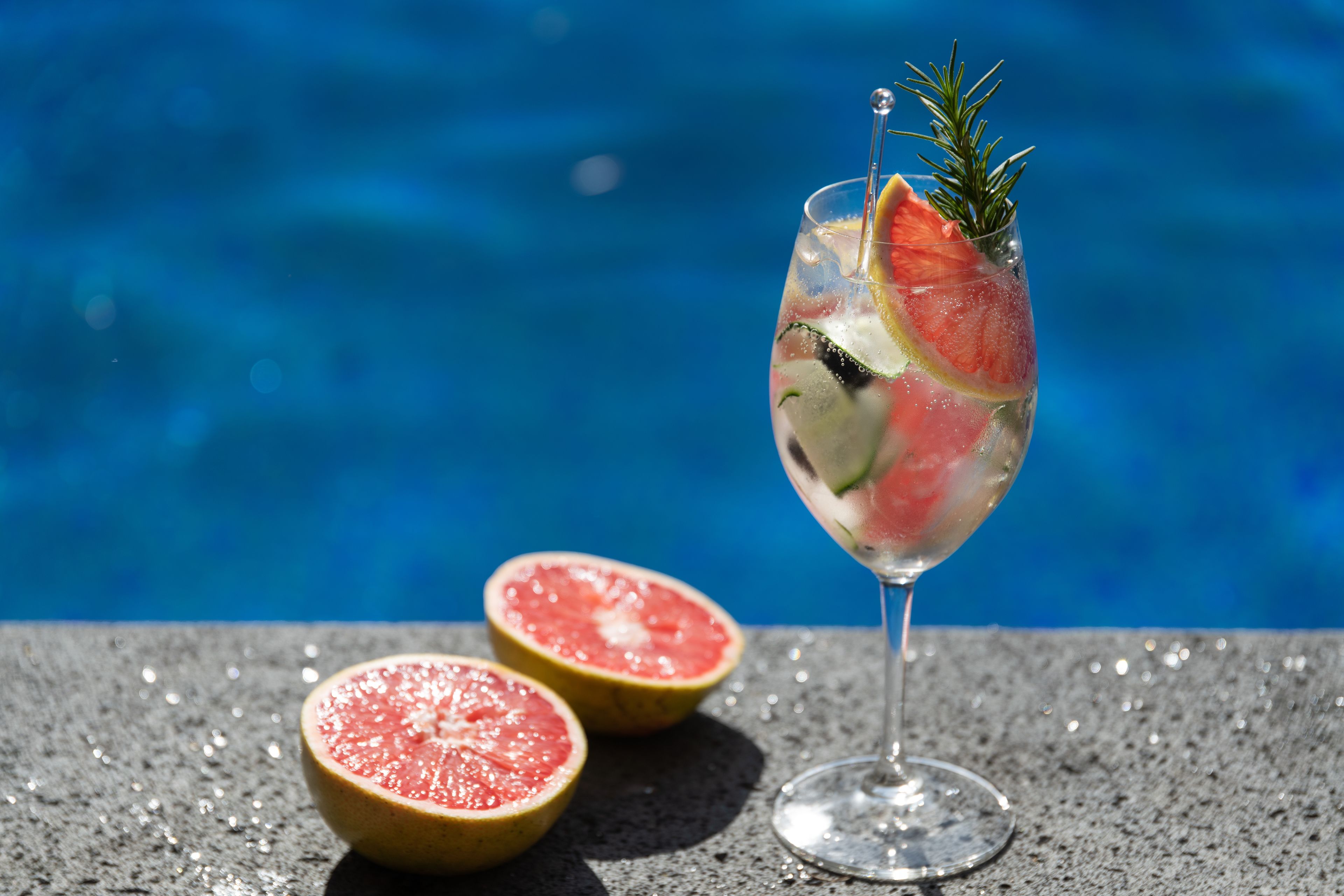 Un verre de cocktail agrémenté d’une tranche de pamplemousse et d’une branche de romarin, à côté d’un pamplemousse entier et d’un demi-pamplemousse, le tout posé sur une surface texturée au bord d’une piscine.