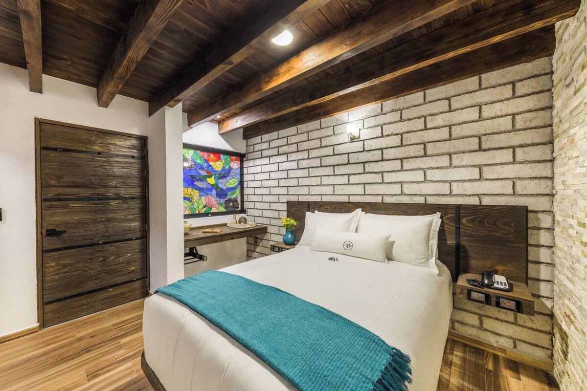A bedroom with a brick wall, wooden ceiling beams, a bed with white bedding, a nightstand with a phone, and a colorful stained glass window.