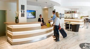 Un espace d’accueil d’hôtel doté d’un comptoir de réception incurvé, d’un écran de télévision et d’un bar avec des places assises.
