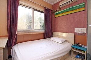 Ein Einzelbett mit weißer Matratze und Kissen, ein Nachttisch mit Lampe und ein Fenster mit Vorhängen.